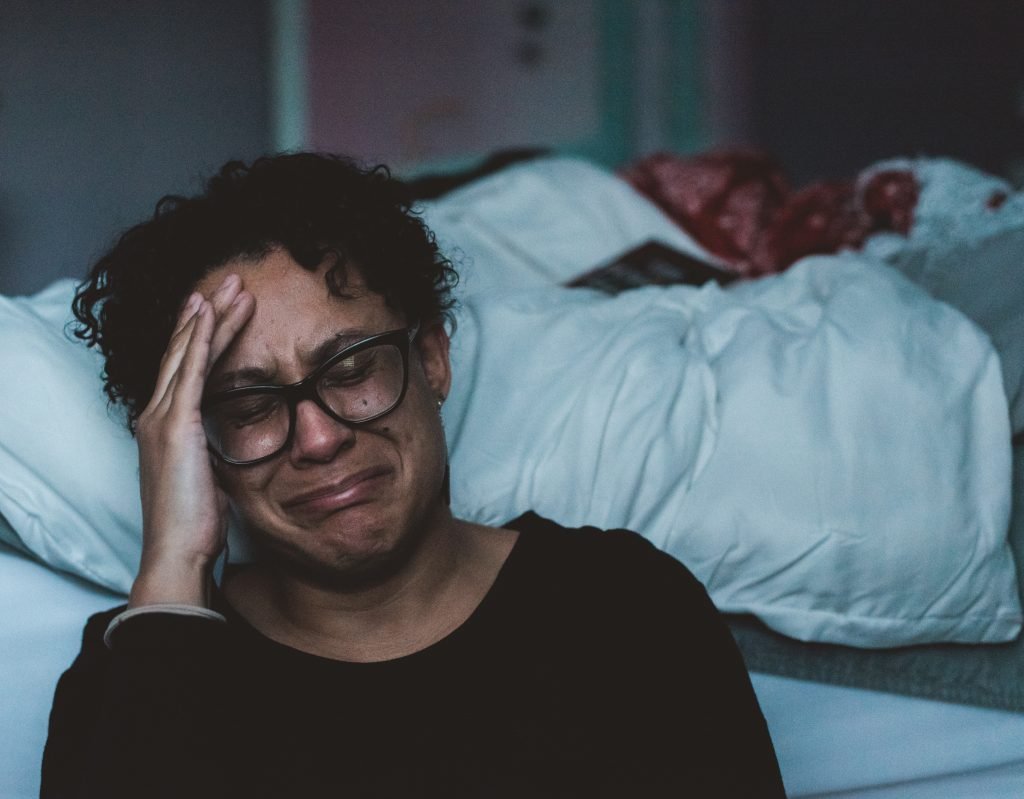 black woman on the floor sitting next to bed crying with hand on her face